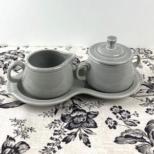 May include: A gray ceramic creamer and sugar bowl set on a white and black floral tablecloth. The creamer and sugar bowl are both in a simple, rounded shape with a smooth, matte finish.