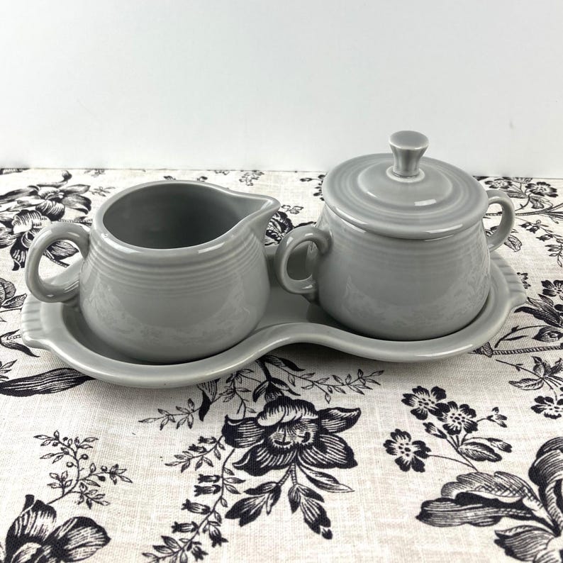 May include: A gray ceramic creamer and sugar bowl set on a white and black floral tablecloth. The creamer and sugar bowl are both in a simple, rounded shape with a smooth, matte finish.