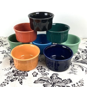 May include: A collection of eight ceramic ramekins in various colors, including black, orange, red, green, blue, and navy. The ramekins are stacked and arranged on a floral patterned surface. These small, round bowls are ideal for baking and serving individual portions.