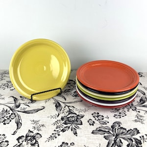 May include: A set of six ceramic dinner plates in various colors, including yellow, orange, white, green, and black. The plates are stacked on top of each other, with the orange plate on top.