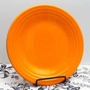 May include: A bright orange ceramic dinner plate with a slightly raised rim. The plate is made by Fiesta and has a smooth, glossy finish.