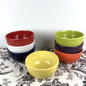May include: A collection of colorful ceramic bowls in various shades, including red, white, navy blue, yellow, green, purple, and orange. The bowls are stacked and arranged on a floral patterned surface.