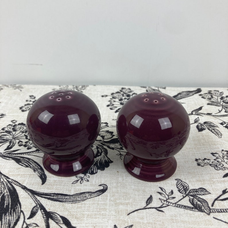 May include: A set of two purple ceramic salt and pepper shakers. The shakers are round with a small base and have a glossy finish.