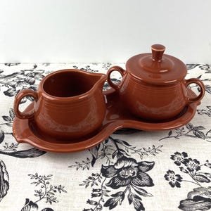 May include: A brown ceramic creamer and sugar bowl set with a matching tray. The set is made in a rustic style with a slightly textured finish.