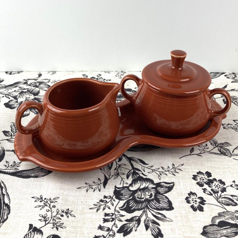 May include: A brown ceramic creamer and sugar bowl set with a matching tray. The set is made in a rustic style with a slightly textured finish.