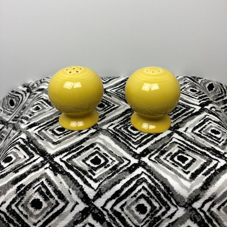 May include: A set of two yellow ceramic salt and pepper shakers. The shakers are round with a small opening on top for dispensing salt and pepper.