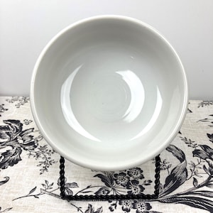 Peut inclure: Un bol rond en c&eacute;ramique blanche avec une finition lisse et brillante et un int&eacute;rieur profond et concave. Le bol est pr&eacute;sent&eacute; sur un support en fil de m&eacute;tal noir torsad&eacute;, offrant une base stable pour voir le r&eacute;cipient circulaire.