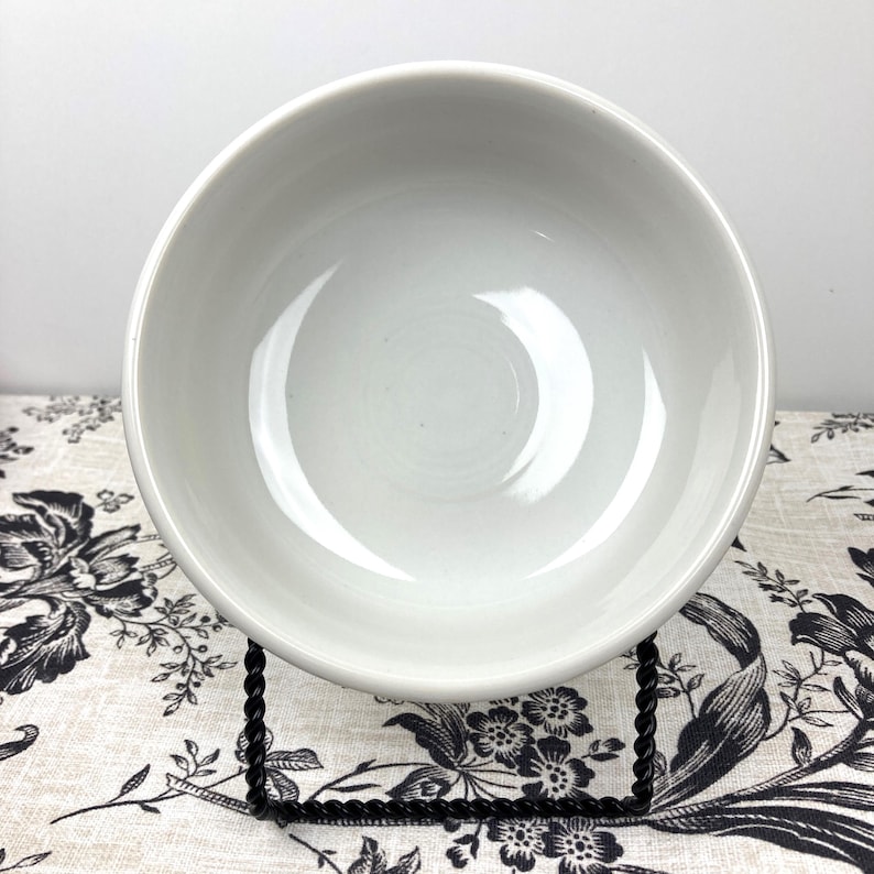 Peut inclure: Un bol rond en c&eacute;ramique blanche avec une finition lisse et brillante et un int&eacute;rieur profond et concave. Le bol est pr&eacute;sent&eacute; sur un support en fil de m&eacute;tal noir torsad&eacute;, offrant une base stable pour voir le r&eacute;cipient circulaire.