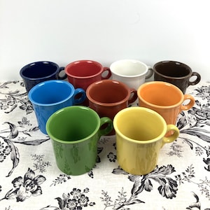 May include: A collection of nine ceramic mugs in various colors, including blue, red, white, brown, orange, green, and yellow. Each mug has a rounded handle and a simple, classic design. The mugs are arranged on a floral patterned tablecloth.