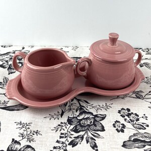 May include: A pink ceramic creamer and sugar bowl set with a matching tray. The set is on a white tablecloth with a black floral pattern.