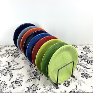 May include: A set of nine round ceramic plates in a spectrum of colors including dark blue, light blue, orange, navy, royal blue, red, yellow-green, forest green, and lime green, arranged vertically in a black metal wire display rack.