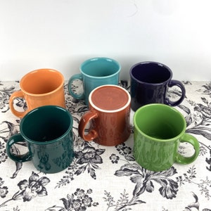 May include: Six ceramic mugs in various colors, including green, blue, orange, brown, and purple. The mugs are arranged in a semi-circle on a floral patterned surface.
