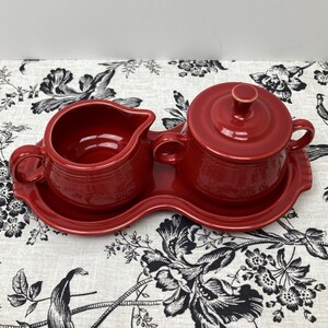 May include: A red ceramic creamer and sugar bowl set with a matching red tray. The set is on a white and black floral tablecloth.