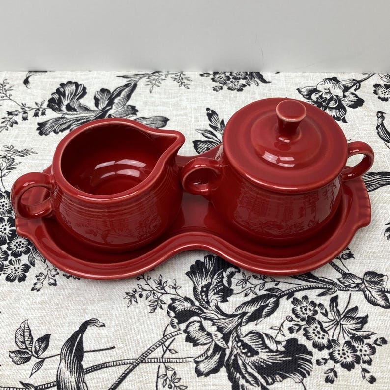 May include: A red ceramic creamer and sugar bowl set with a matching red tray. The set is on a white and black floral tablecloth.