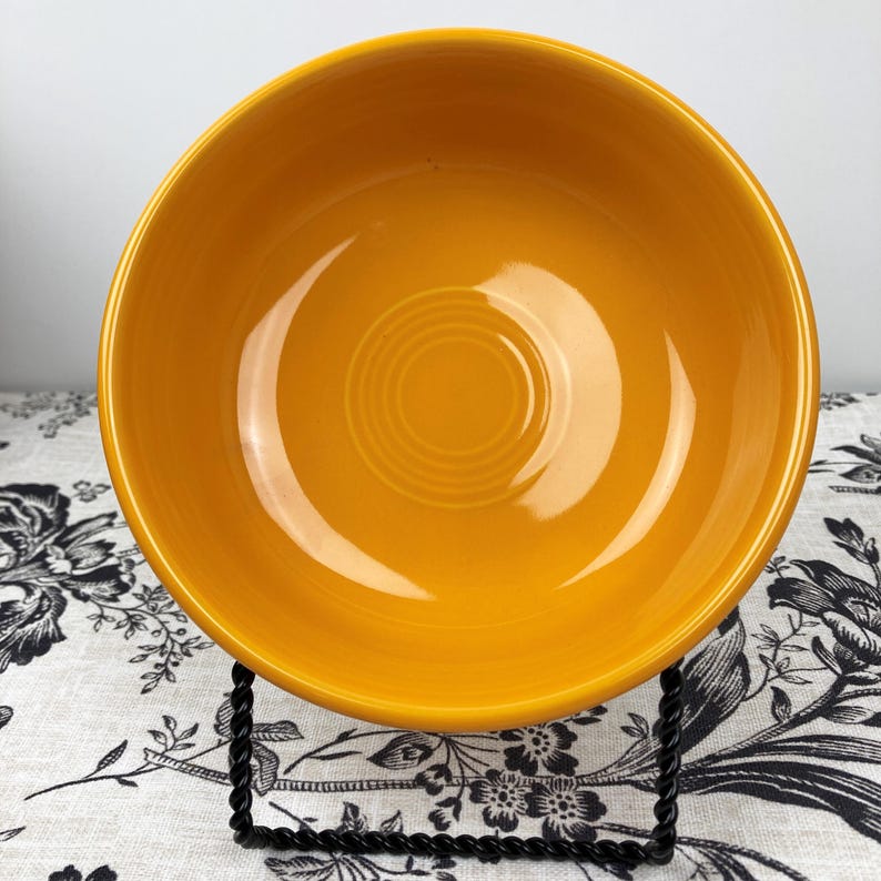 Peut inclure: Un bol en c&eacute;ramique jaune moutarde brillant avec un int&eacute;rieur lisse et arrondi, pr&eacute;sentant une s&eacute;rie de cercles concentriques en relief &agrave; la base. Le bol est pos&eacute; sur un support en fil m&eacute;tallique noir, r&eacute;v&eacute;lant son bassin profond.