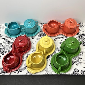 May include: Set of five colorful ceramic creamer and sugar bowls with matching serving trays. Each set features a different color: red, blue, green, yellow, and orange.
