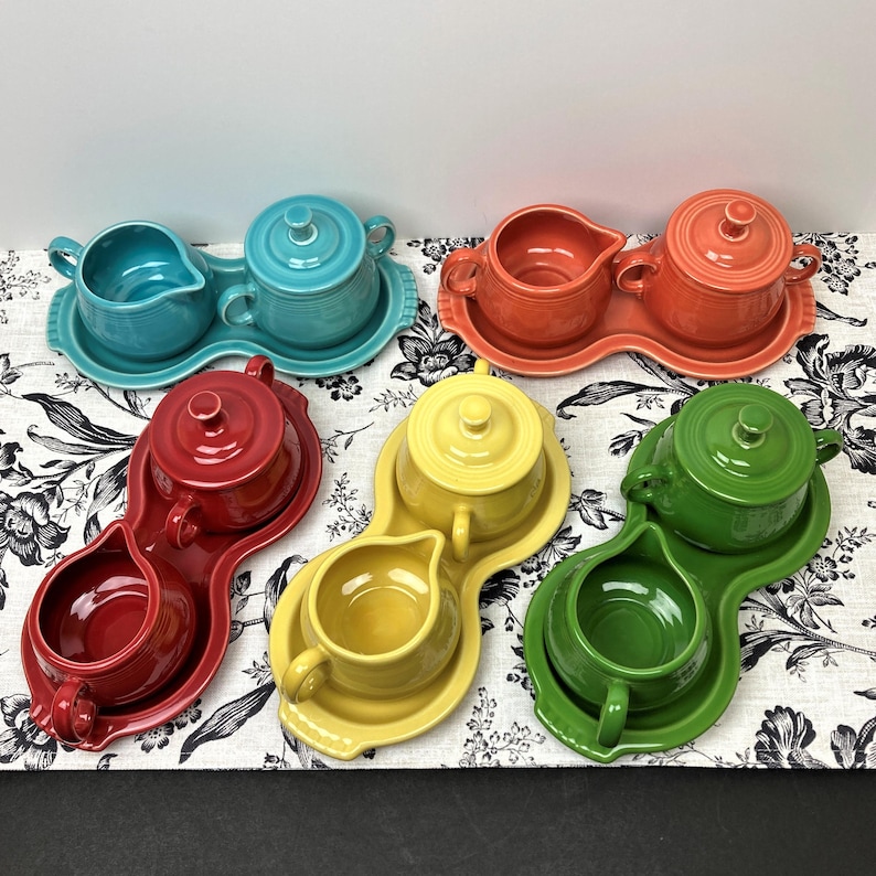 May include: Set of five colorful ceramic creamer and sugar bowls with matching serving trays. Each set features a different color: red, blue, green, yellow, and orange.