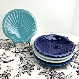 May include: A set of five ceramic plates shaped like seashells, featuring a glossy finish. One turquoise plate stands upright on a black wire display stand, while four additional plates in dark blue, light blue, white, and navy are stacked below.