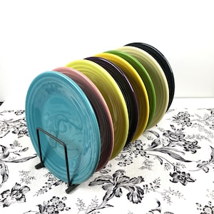 May include: A set of seven colorful ceramic plates stacked on a black metal plate stand. The plates are in shades of blue, green, yellow, pink, and purple.
