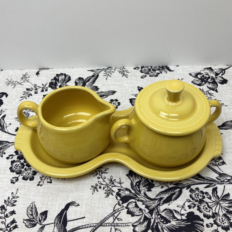 May include: A yellow ceramic creamer and sugar bowl set with a matching tray. The set is on a floral tablecloth.