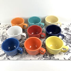 May include: A collection of eight ceramic mugs in various colors, including orange, teal, yellow, white, red, and navy blue. Each mug has a rounded shape with a handle. The mugs are arranged on a floral patterned fabric.