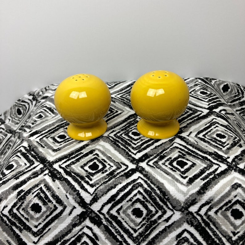 May include: A set of two yellow ceramic salt and pepper shakers. The shakers are round with a small opening on top for dispensing salt and pepper. They are sitting on a black and white patterned surface.