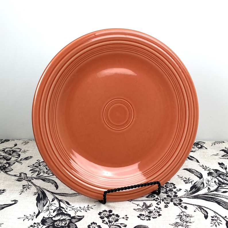 Pu&ograve; includere: Un piatto da portata di colore arancio corallo con un bordo rialzato e cerchi concentrici al centro. Il piatto &egrave; in ceramica e ha una finitura lucida.