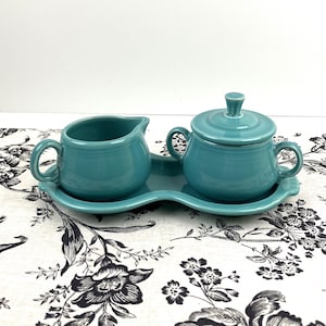May include: A teal ceramic creamer and a lidded sugar bowl set resting on a matching oval tray. The creamer features a curved handle and spout, while the sugar bowl has two side handles and a knobbed lid, all with a smooth, glossy finish.
