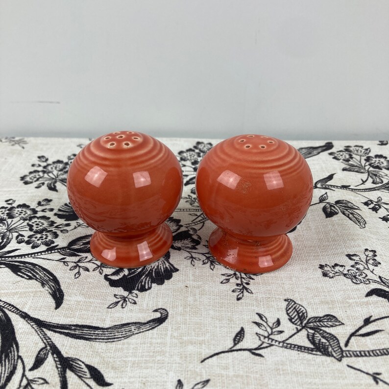 May include: A pair of salt and pepper shakers in a vibrant orange colour. The shakers are round with a textured surface and a small opening on top for dispensing salt and pepper.
