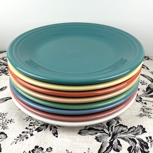 May include: A stack of seven ceramic dinner plates in various pastel colors, including teal, yellow, orange, pink, green, blue, and white.