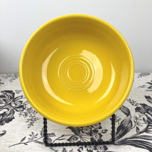 Peut inclure: Un bol en c&eacute;ramique jaune vif avec une finition lisse et brillante, pr&eacute;sentant une s&eacute;rie de cercles concentriques en relief au centre de la base int&eacute;rieure. Le bol poss&egrave;de un bord large et arrondi et est pos&eacute; sur un support en fil noir.