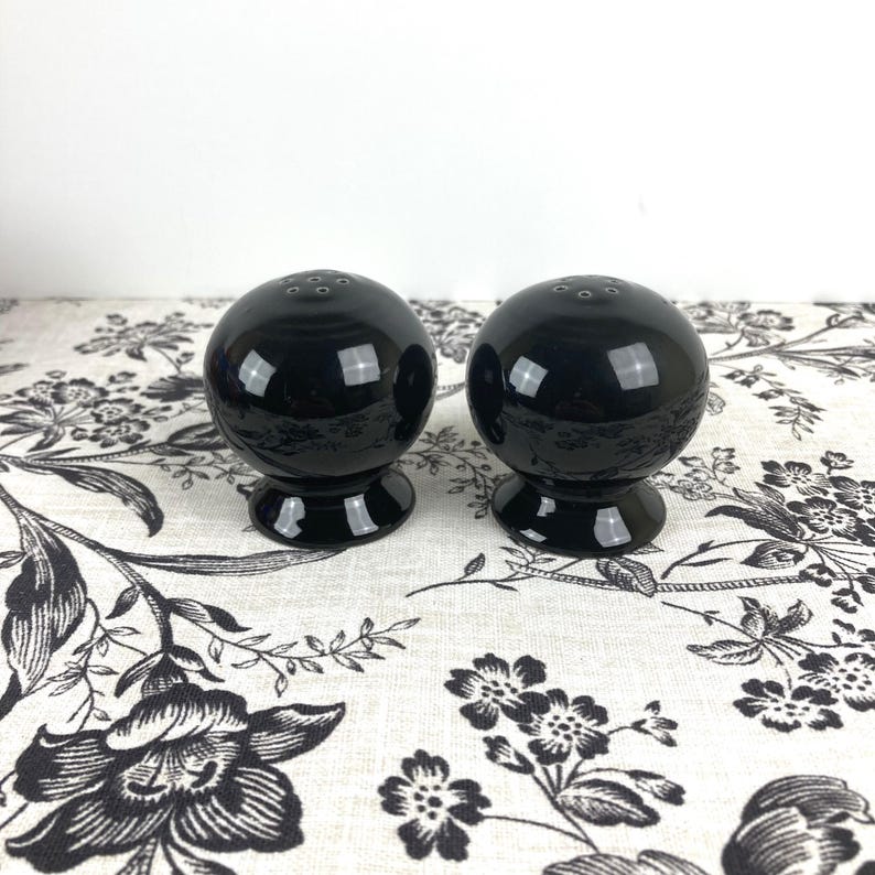 May include: Two black ceramic salt and pepper shakers with a round, smooth design. The shakers are sitting on a white tablecloth with a black floral pattern.