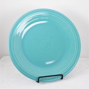Pu&ograve; includere: Un piatto da portata in ceramica blu turchese con un bordo rialzato e un design sottile di cerchi concentrici. Il piatto &egrave; su un supporto nero.