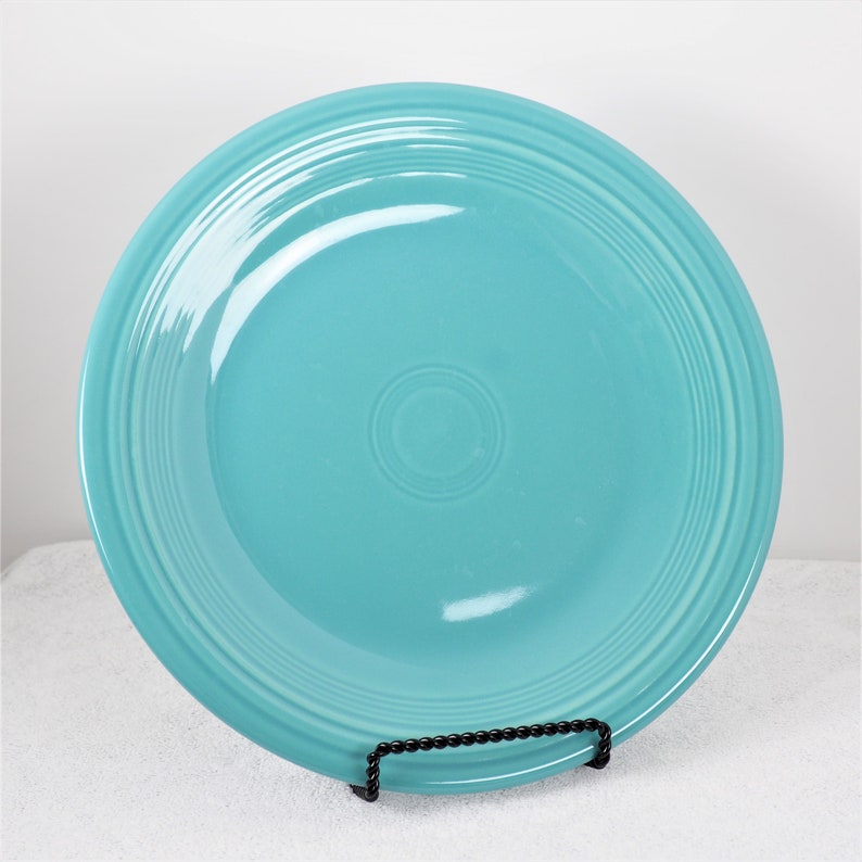 Pu&ograve; includere: Un piatto da portata in ceramica blu turchese con un bordo rialzato e un design sottile di cerchi concentrici. Il piatto &egrave; su un supporto nero.