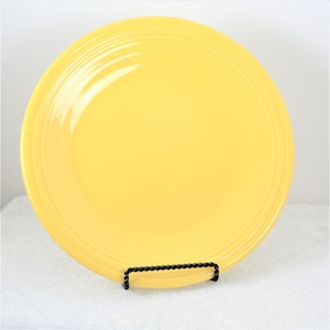 Pu&ograve; includere: Un piatto da cena in ceramica giallo brillante con un bordo leggermente rialzato e un design sottile di cerchi concentrici. Il piatto &egrave; su un supporto in metallo nero.