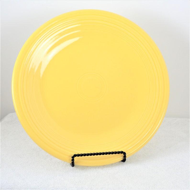 Pu&ograve; includere: Un piatto da cena in ceramica giallo brillante con un bordo leggermente rialzato e un design sottile di cerchi concentrici. Il piatto &egrave; su un supporto in metallo nero.