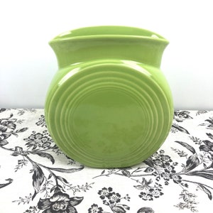 May include: A bright green ceramic vase with a rounded body and a flared rim. The vase features concentric circle detailing on the front. The vase is sitting on a floral patterned cloth.