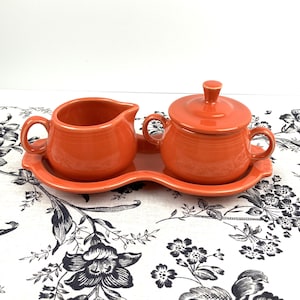 May include: A bright orange ceramic creamer and a lidded sugar bowl with two handles, both resting on a matching orange oval tray with a scalloped edge. The set features a smooth, glossy finish and concentric ring detailing on the exterior surfaces.