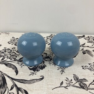 May include: A pair of blue ceramic salt and pepper shakers. The shakers are round with a small base and have holes on the top for dispensing salt and pepper.