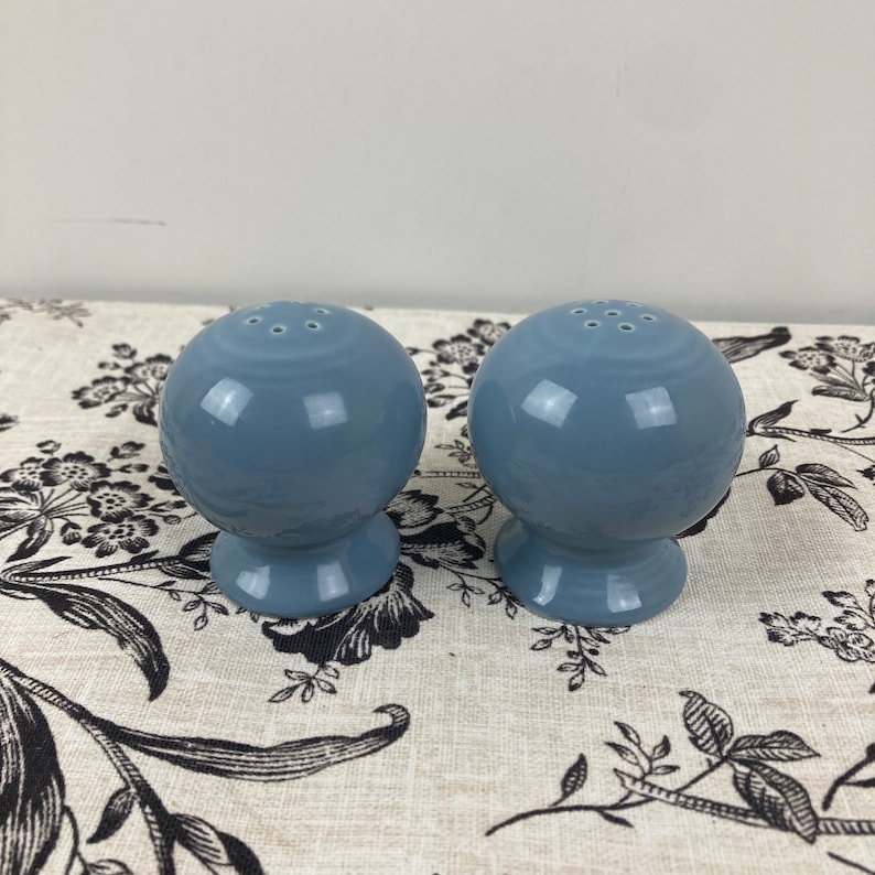 May include: A pair of blue ceramic salt and pepper shakers. The shakers are round with a small base and have holes on the top for dispensing salt and pepper.