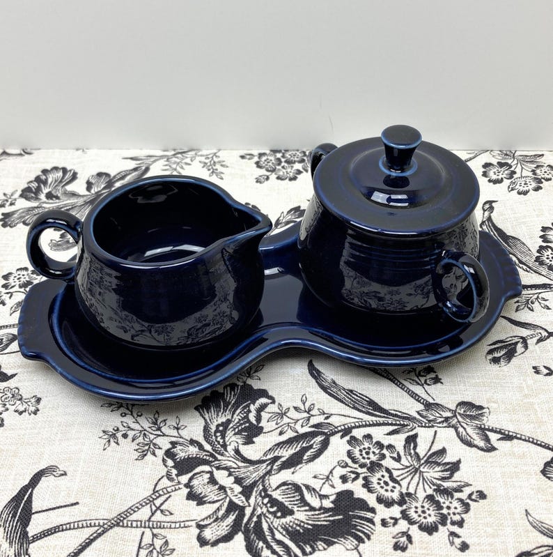 May include: A blue ceramic creamer and sugar bowl set with a matching tray. The set is in a deep blue color and has a simple, elegant design.