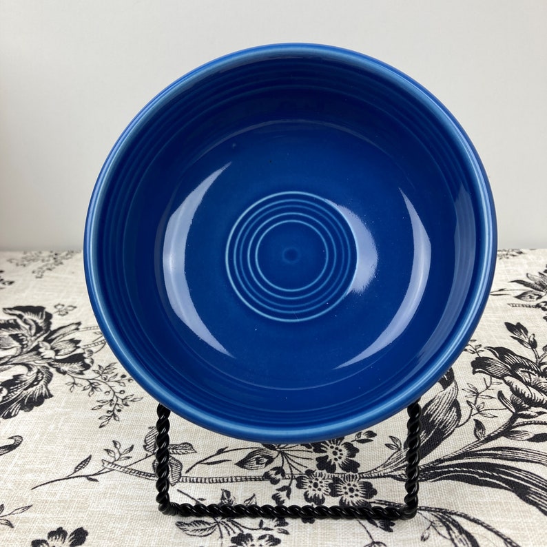 Peut inclure: Un bol en c&eacute;ramique bleu cobalt profond avec une finition lisse et brillante, pr&eacute;sentant une s&eacute;rie de cercles concentriques en relief au fond. Le bol est pr&eacute;sent&eacute; sur un support en fil m&eacute;tallique noir, mettant en valeur sa forme ronde.