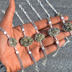 May include: Several silver bracelets with pyrite stones are displayed. Each bracelet features a unique, irregularly shaped pyrite stone set in a silver bezel. The bracelets have a silver chain and are shown on a hand.