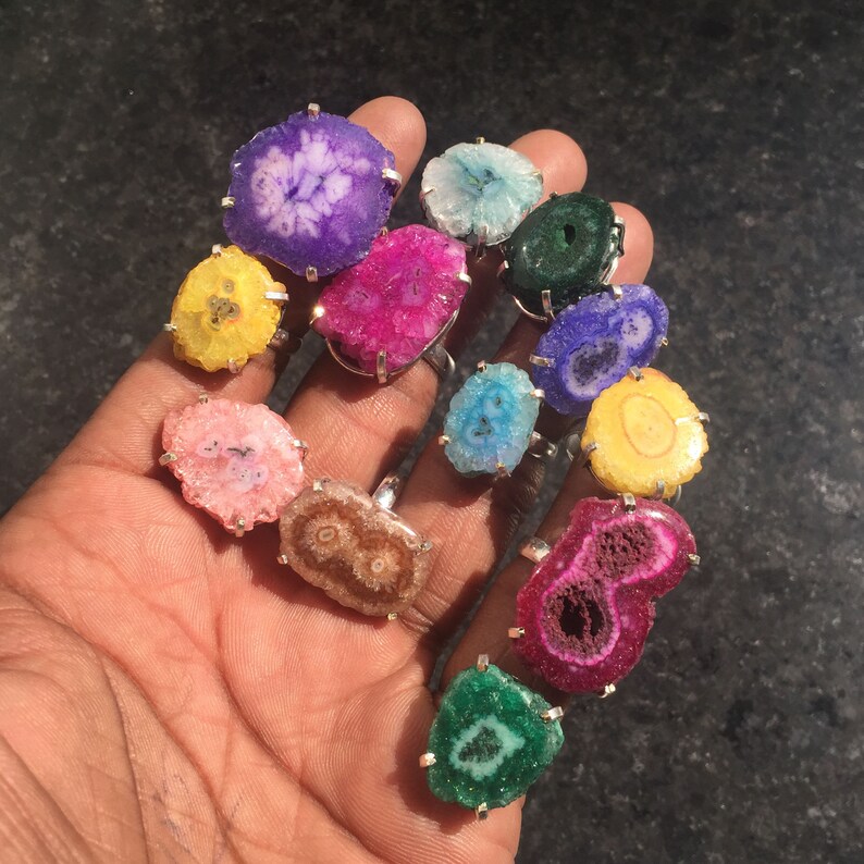 Pu&ograve; includere: Una collezione di 11 anelli con fette di agata colorate incastonate in argento. Gli anelli sono disposti su una mano, mostrando i motivi e i colori unici di ogni pietra.