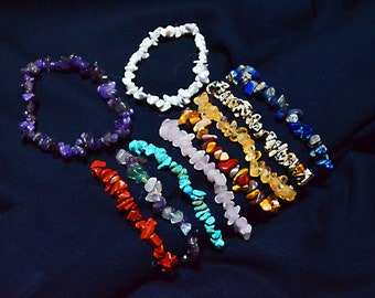 Puede incluir: Un conjunto de diez pulseras hechas con varias piedras semipreciosas, incluyendo amatista, turquesa, cuarzo rosa y ojo de tigre. Las pulseras están dispuestas en un patrón de arcoíris sobre un fondo de tela azul oscuro.