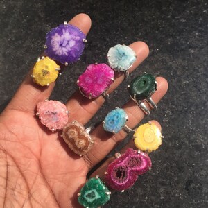 Pu&ograve; includere: Una collezione di 12 anelli con fette di agata colorate incastonate in argento. Ogni anello ha un motivo e un colore unici, tra cui viola, rosa, giallo, verde e blu.