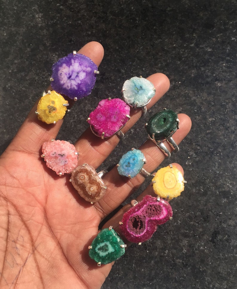 Pu&ograve; includere: Una collezione di 12 anelli con fette di agata colorate incastonate in argento. Ogni anello ha un motivo e un colore unici, tra cui viola, rosa, giallo, verde e blu.