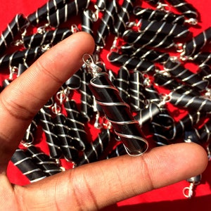 Puede incluir: Primer plano de una mano sosteniendo un colgante de obsidiana negra envuelto en alambre de plata. El colgante es un cilindro largo y delgado con un bucle de plata en la parte superior. El colgante est&aacute; rodeado de otros colgantes similares.
