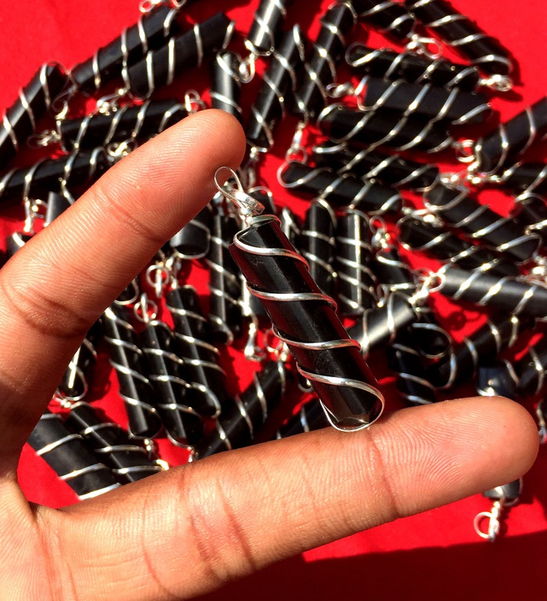 Puede incluir: Primer plano de una mano sosteniendo un colgante de obsidiana negra envuelto en alambre de plata. El colgante es un cilindro largo y delgado con un bucle de plata en la parte superior. El colgante est&aacute; rodeado de otros colgantes similares.