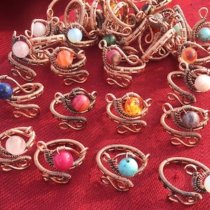May include: A selection of handcrafted copper wire rings, each with a different gemstone. The rings have an intricate spiral design, and the gemstones vary in colour, including pink, blue, orange, and white. The rings are displayed on a red surface.
